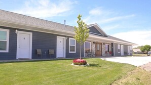 A two-story building with a grey exterior, numbered doors, and a small garden with red flowers. - Greystone Crossing Suite #11 (Heyburn)