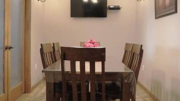 A dining area with a rectangular table, six chairs, a chandelier, wall-mounted lights, a flat-screen TV, and a framed picture on the wall.