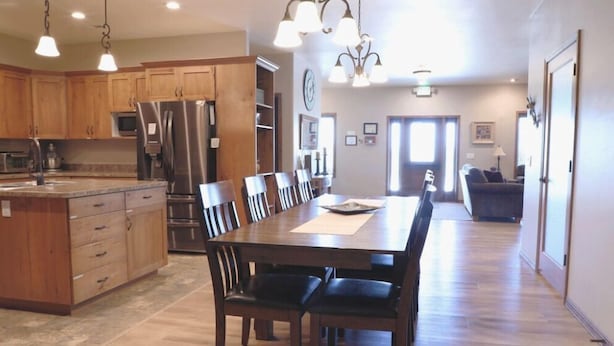 A spacious kitchen with wooden cabinets, a stainless steel refrigerator, and a dining area with a long table and chairs. - Greystone Crossing Suite #11 (Heyburn)