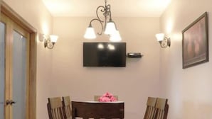 A dining area with a wooden table and chairs, a chandelier, wall-mounted lights, and a television on the wall. - Greystone Crossing Suite #12 (Heyburn)