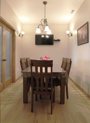 A dining area with a wooden table and chairs, a chandelier, wall-mounted lights, and a television on the wall. - Greystone Crossing Suite #12 (Heyburn)