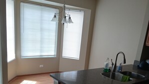 A kitchen with a granite countertop, a stainless steel sink, and a black trash can. There are two windows with blinds, and a chandelier hangs above the counter.
