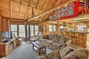 L’intérieur chaleureux d’un chalet avec un grand plafond en bois, un espace de vie comprenant un canapé et des fauteuils, un téléviseur et un coin cuisine avec des tabourets de bar.
