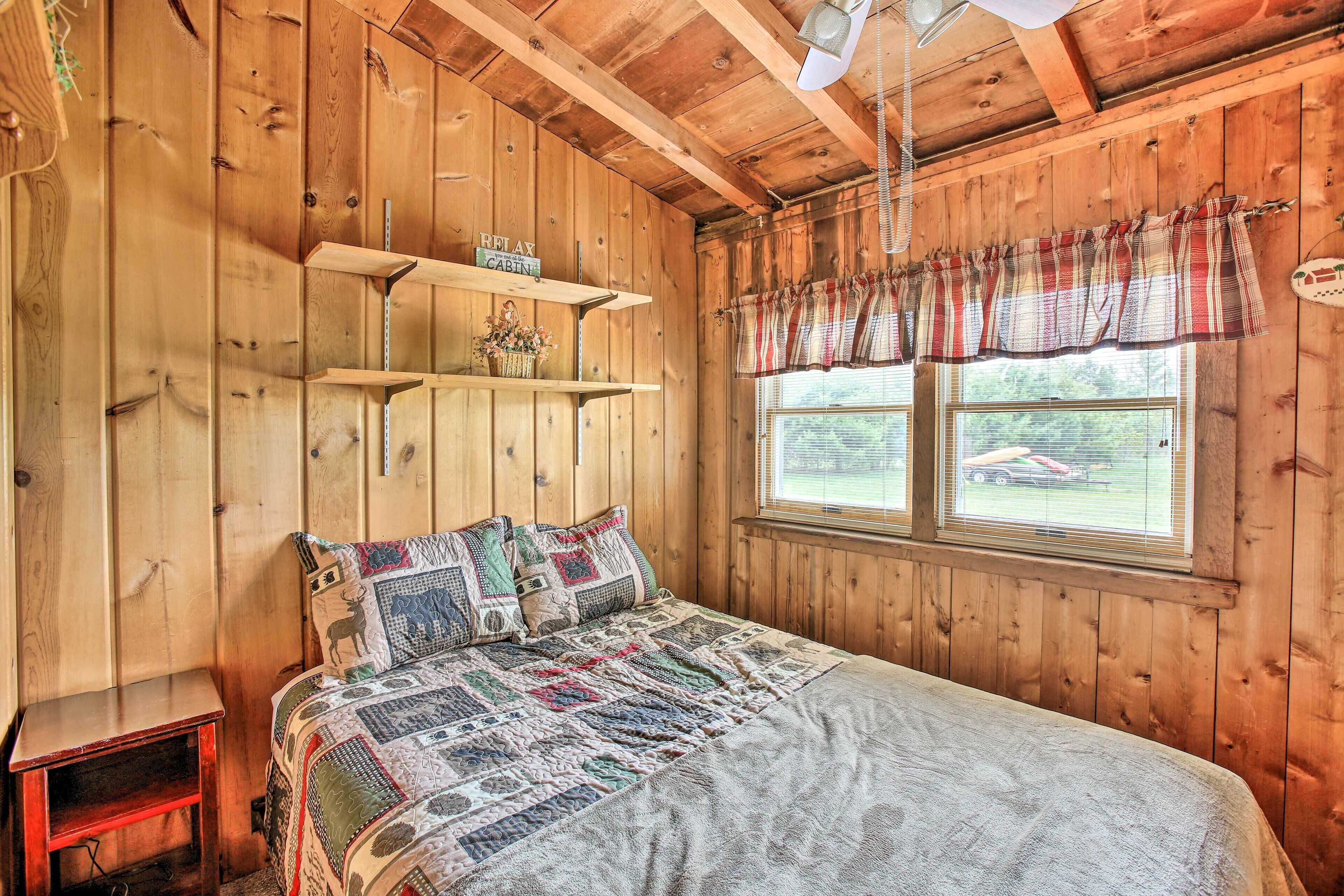 A wooden cabin room with a bed, shelves, and a window with a plaid valance.