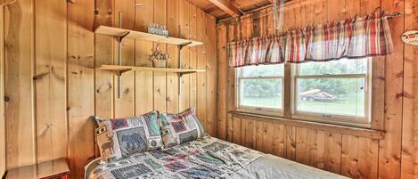 A wooden cabin room with a bed, shelves, and a window with a plaid valance.