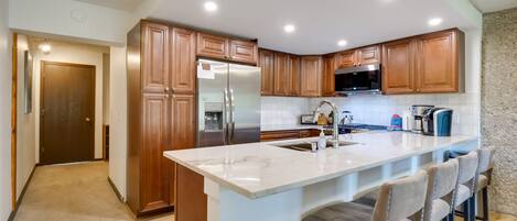 A modern kitchen with wooden cabinets, a stainless steel refrigerator, a microwave, and a marble island with bar stools.