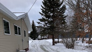 Une allée enneigée menant à une maison sur laquelle on peut voir un climatiseur et où se trouve un petit arbre dans le jardin.
