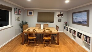 Un coin repas avec une table et des chaises en bois, une fenêtre avec des stores, des tableaux encadrés aux murs et une bibliothèque remplie de livres.