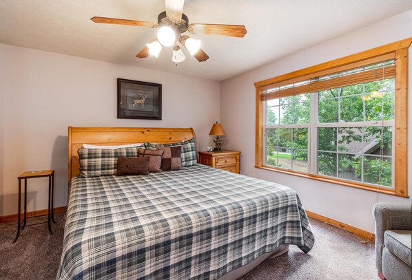 A bedroom with a large bed, a ceiling fan, a window with a view of greenery, and a framed picture on the wall. - Bear Cabin Getaway (Reeds Spring)