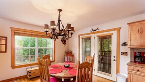 A dining area with a wooden table, four chairs, a chandelier, and large windows with blinds. - Bear Cabin Getaway (Reeds Spring)