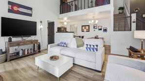 Een ruime woonkamer met een witte bank, een salontafel, een aan een houten console bevestigde televisie, en een trap naar een hoger gelegen verdieping.
