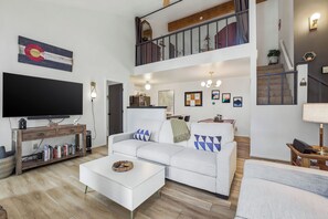 Ein großzügiger Wohnbereich mit einem weißen Sofa, einem Couchtisch, einem an einer Holzkonsolenwand montierten Fernseher und einer Treppe, die in ein Obergeschoss führt.