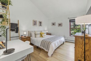 A bedroom with a bed, bedside tables, a desk, a lamp, a plant shelf, and a window.