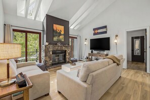 A modern living room with a stone fireplace, a flat-screen TV, and a large window overlooking greenery.