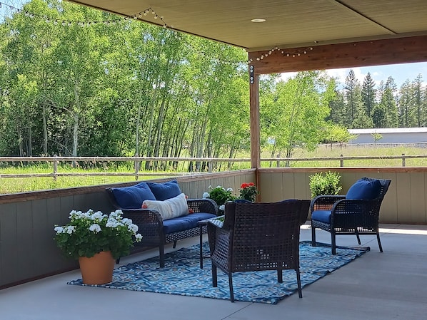 Une terrasse couverte, meublée avec des meubles en osier, agrémentée d’une plante en pot et offrant une vue sur une zone boisée.