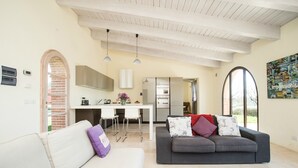 A modern living room with a grey sofa and a white couch, a kitchen in the background, and a large window providing natural light. - Villino Rosmarino - Cignella Resort Toscana (Toscana)