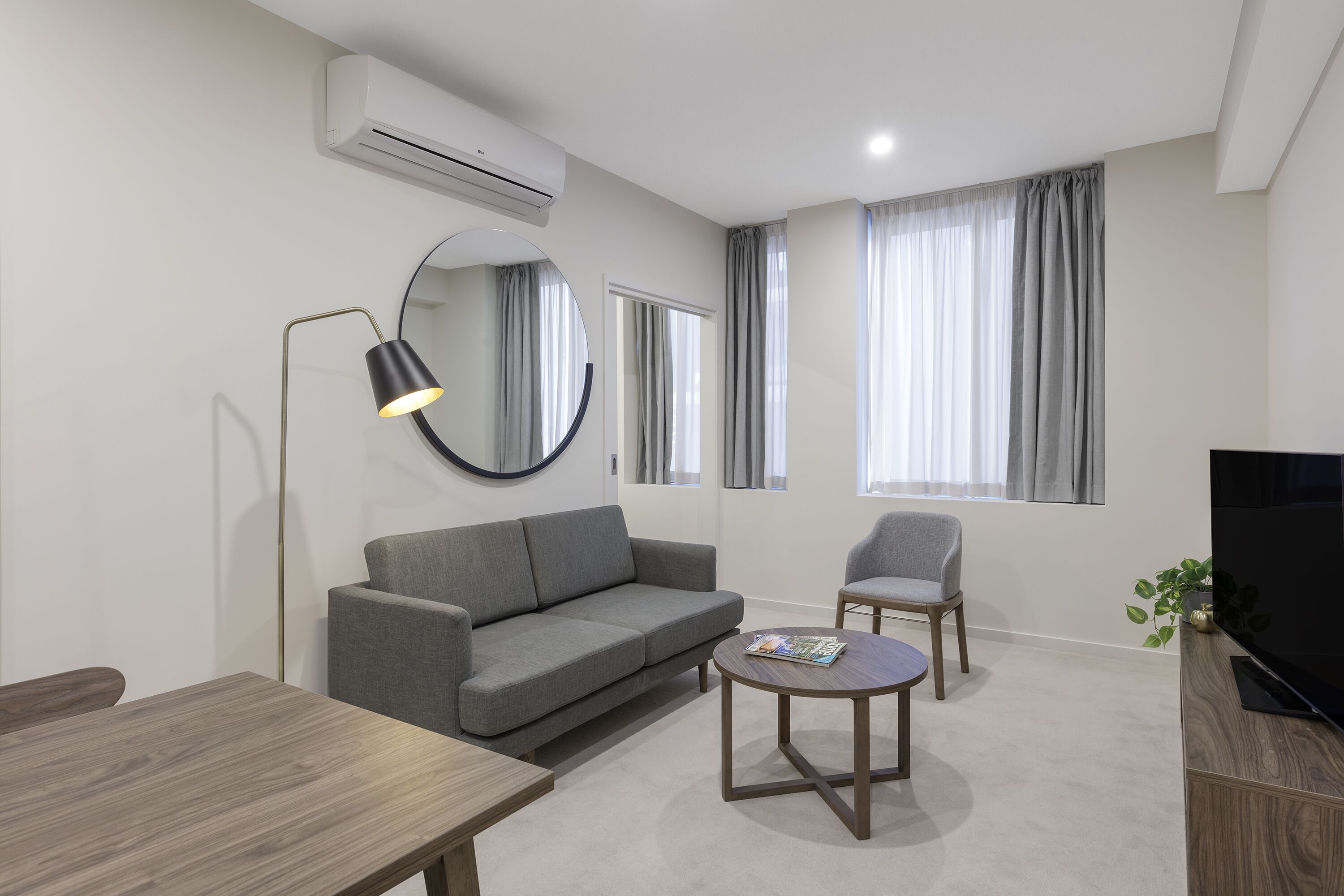 Un salon moderne avec un canapé gris, un miroir rond, une petite table, une chaise et un téléviseur posé sur un meuble en bois.