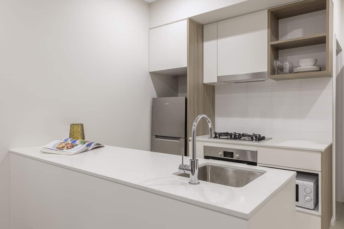 Una cocina moderna con armarios blancos, un lavamanos de acero inoxidable, un horno empotrado y una pequeña estantería con cristalería.