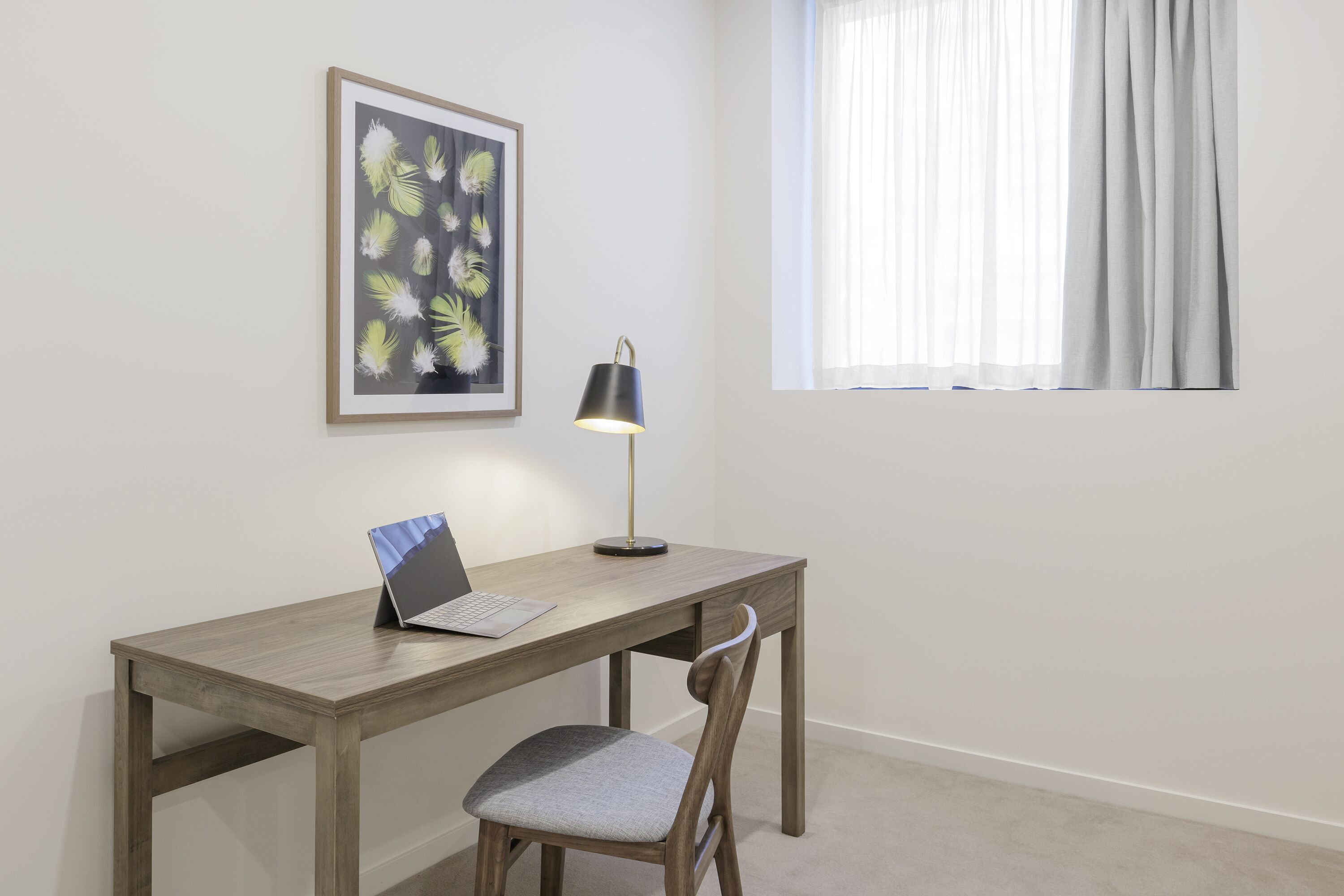 A room with a wooden desk, a laptop, a chair, a desk lamp, and a framed picture on the wall.