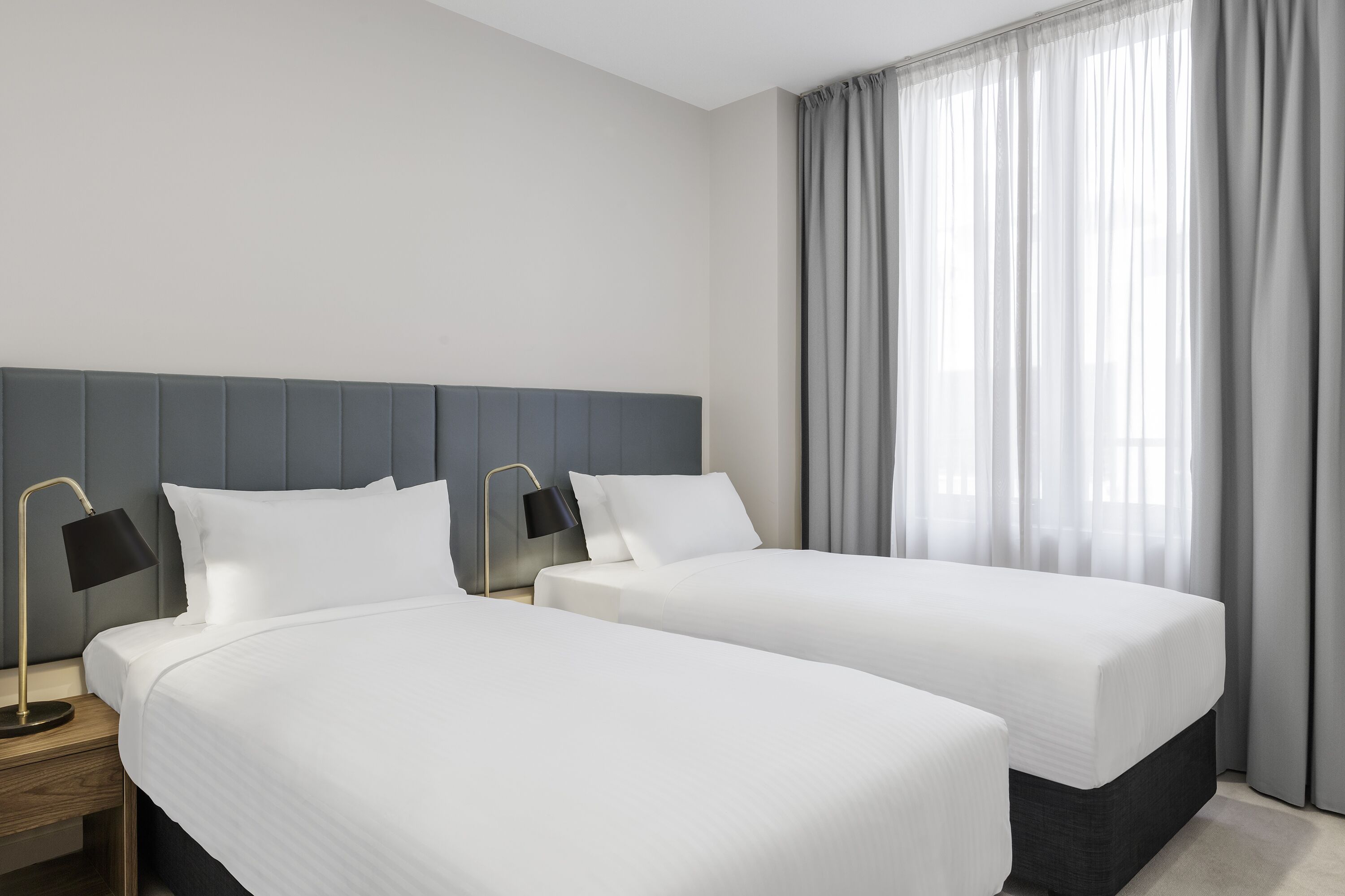 A hotel room with two single beds, a nightstand with a lamp, and a large window with curtains.