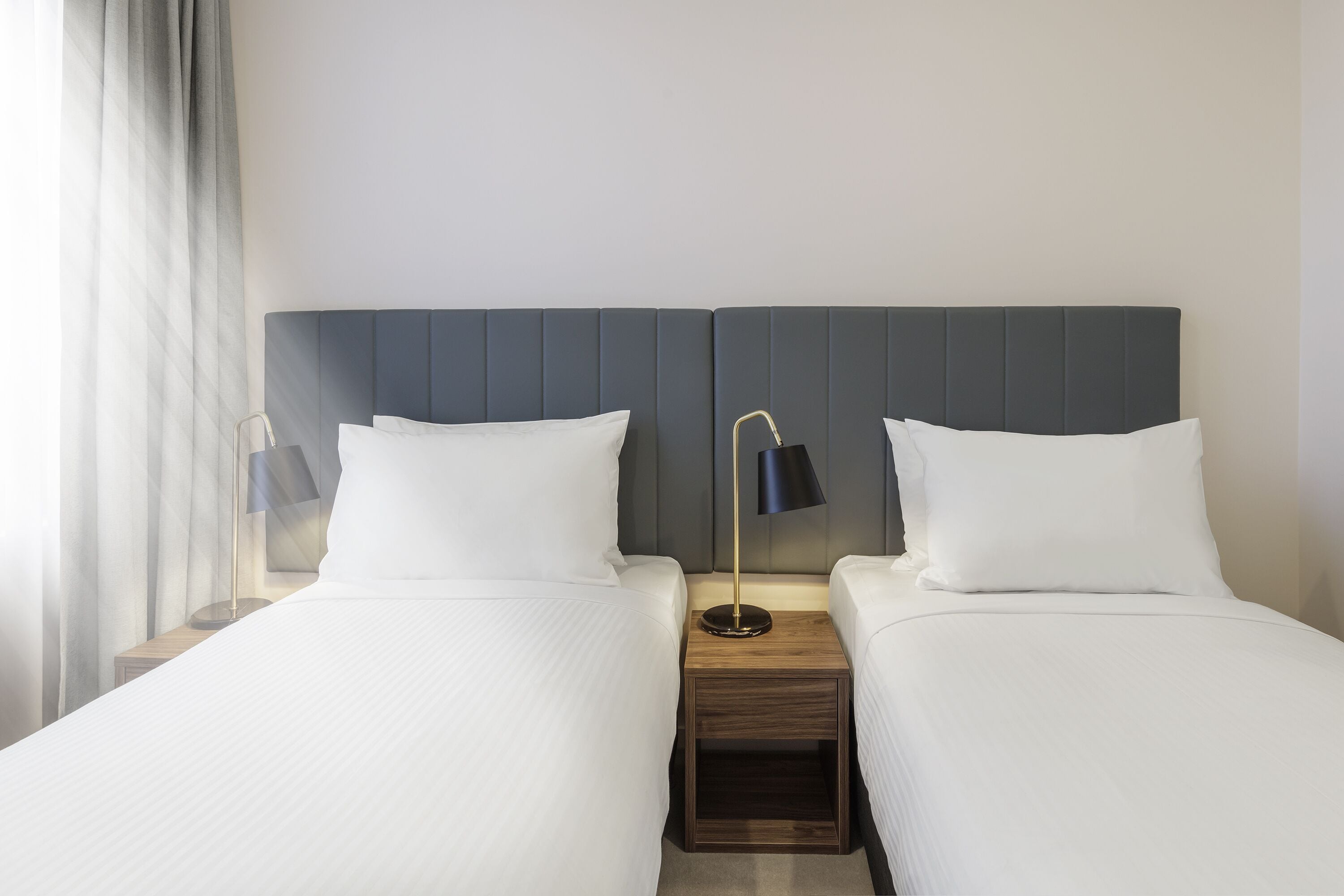 A hotel room with two beds, a nightstand with a lamp, and a window with curtains.