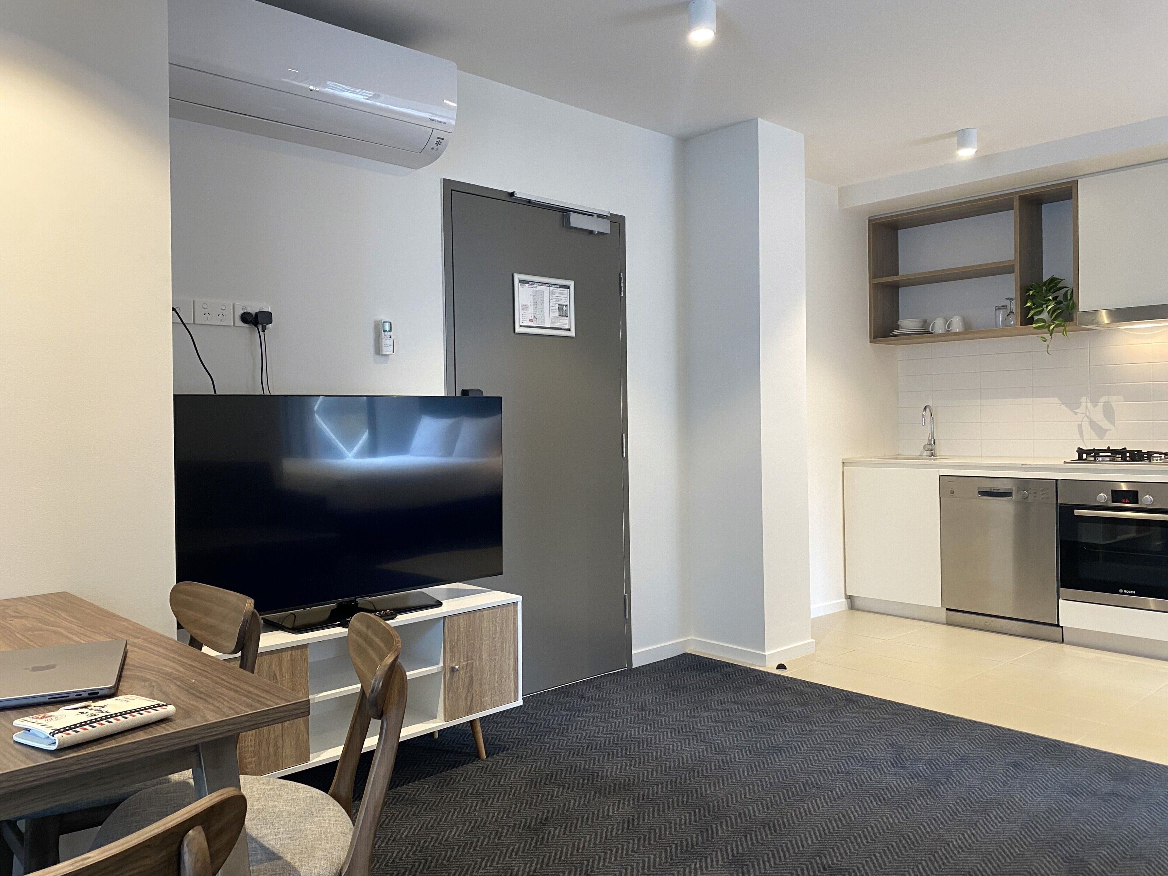A modern living room with a flat-screen TV, a dining area with a laptop and remote, a kitchen with a dishwasher and stove, and a gray herringbone-patterned carpet.