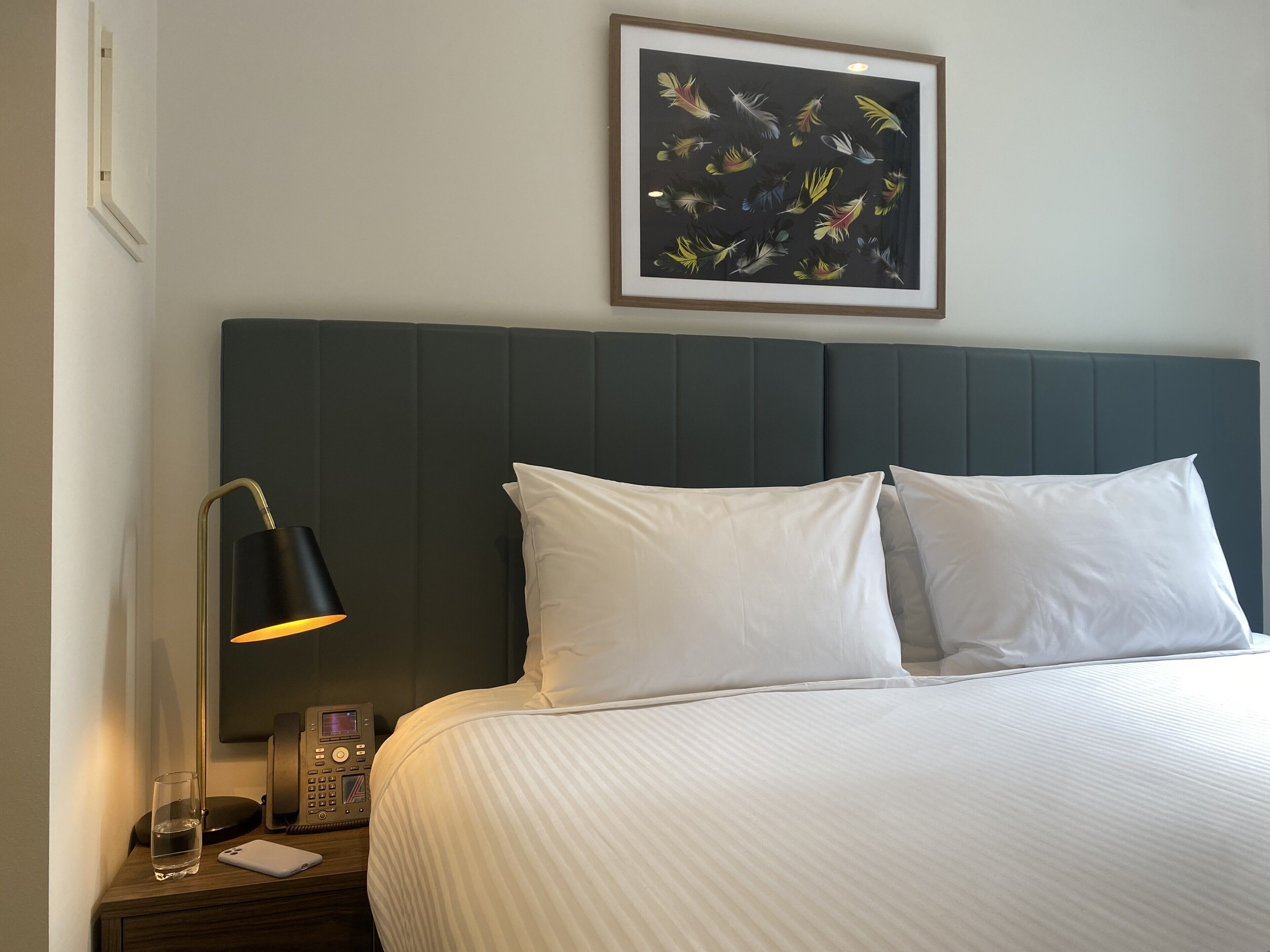 A neatly made bed with white linens, a dark headboard, a bedside table with a lamp, a glass of water, and a phone.