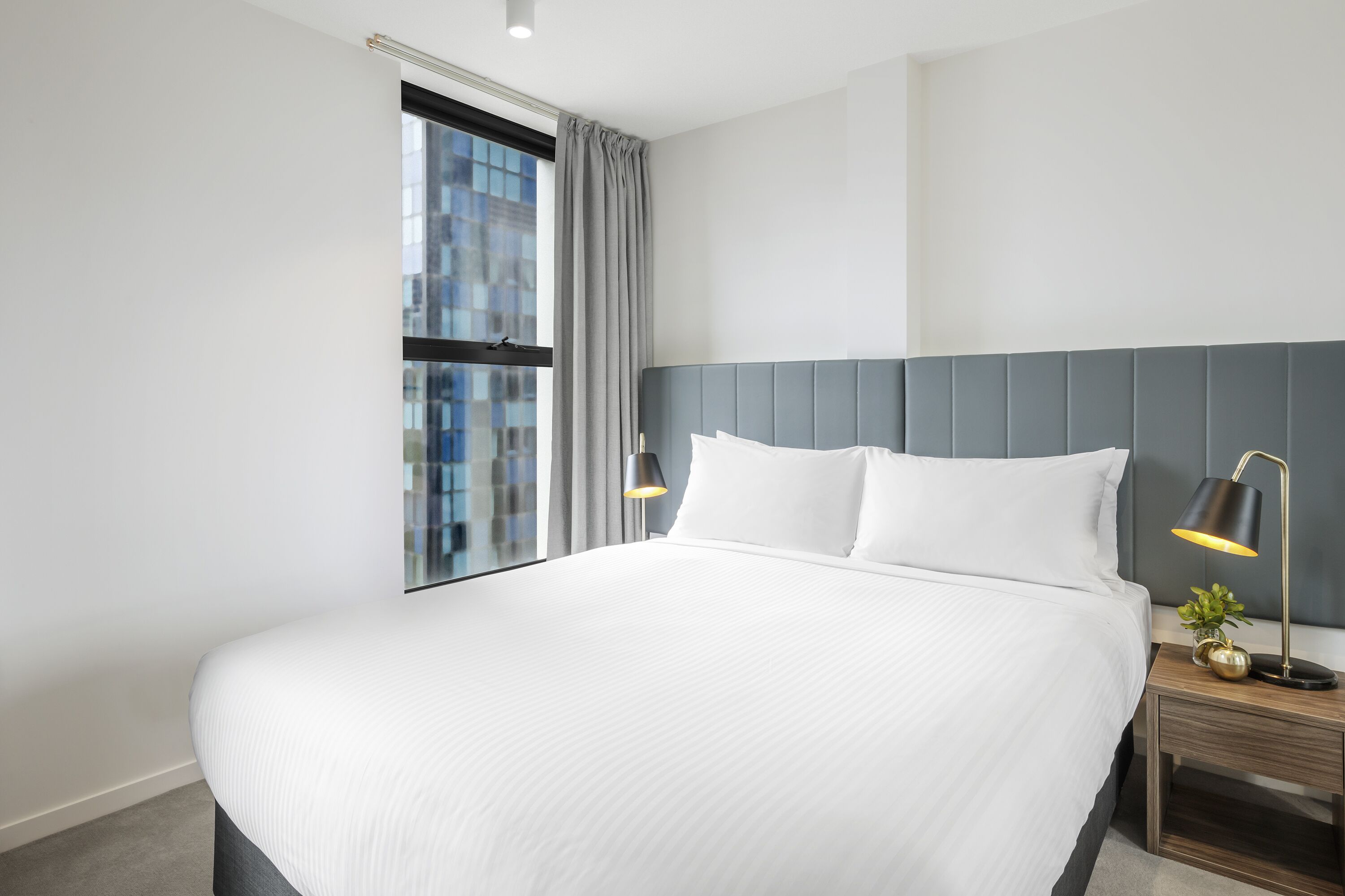 A modern hotel room with a large bed, a nightstand with a lamp, and a window with curtains.
