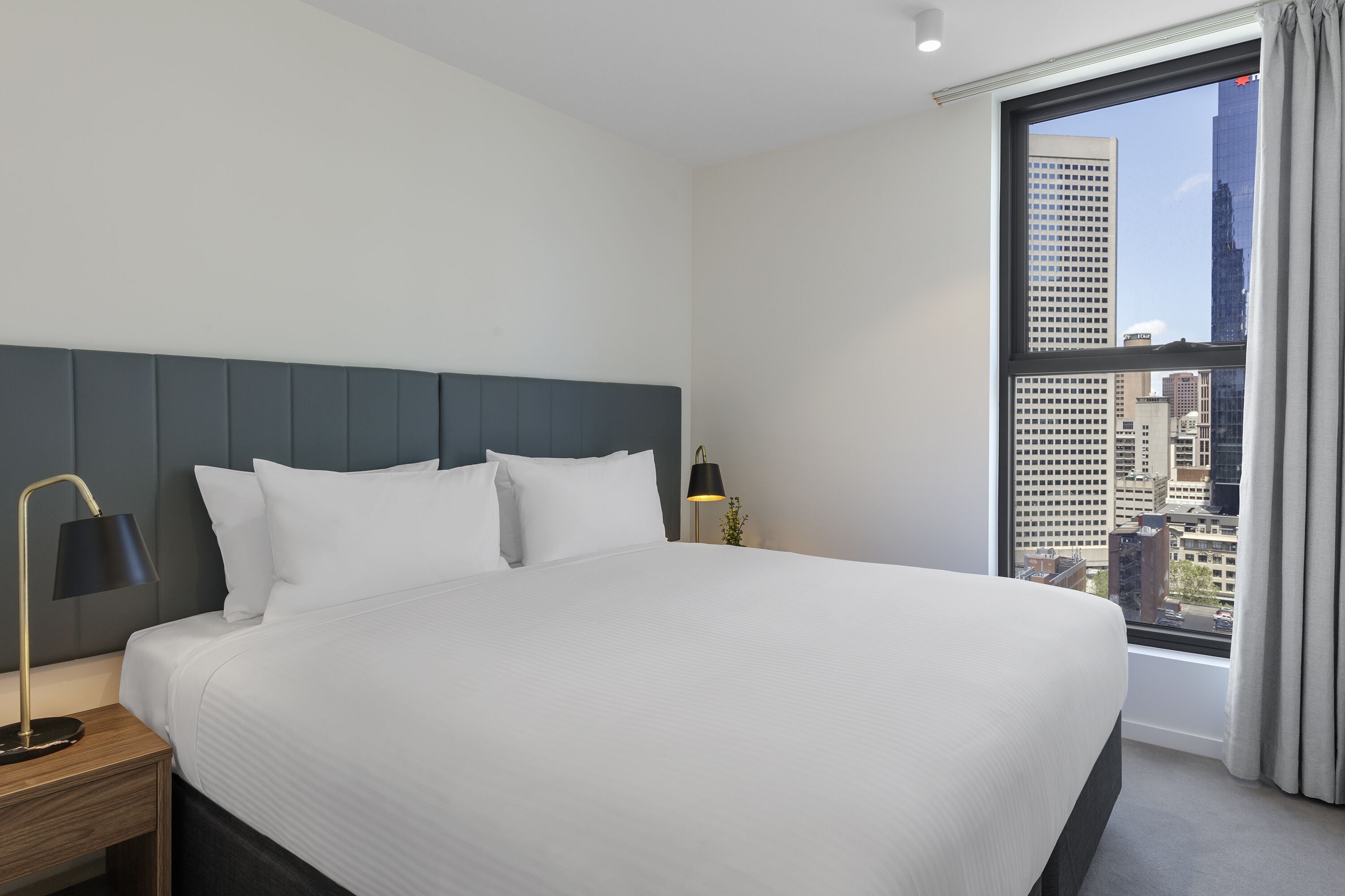 A hotel room with a large bed, a nightstand with a lamp, and a view of a cityscape through a window.
