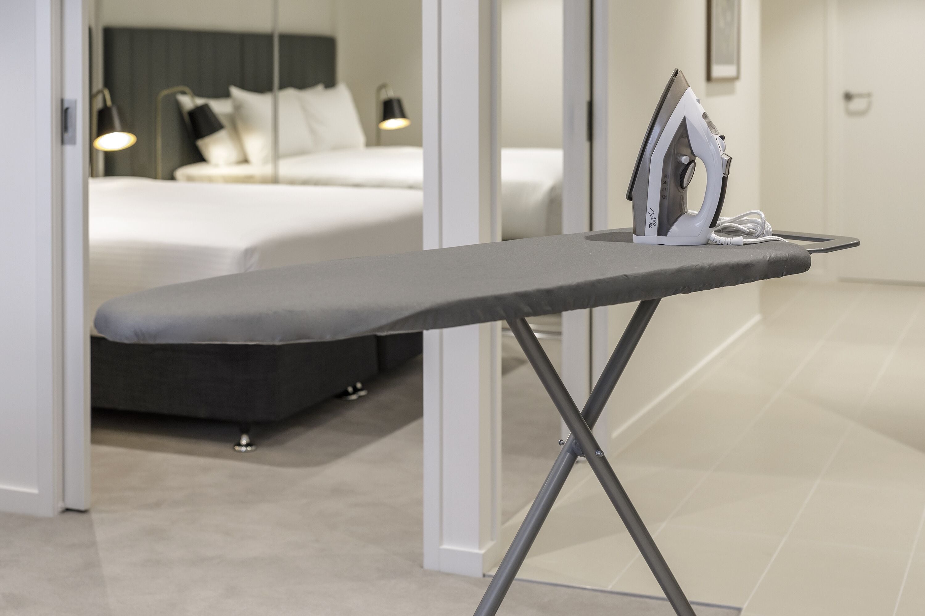 Habitación de hotel moderna con una cama bien hecha, una lámpara de noche y una tabla de planchar con plancha y electrodoméstico conectado.