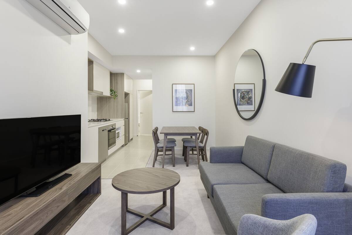 Una sala de estar moderna con un televisor de pantalla plana, un sofá gris, una mesa de centro redonda y una zona de comedor con mesa y sillas.