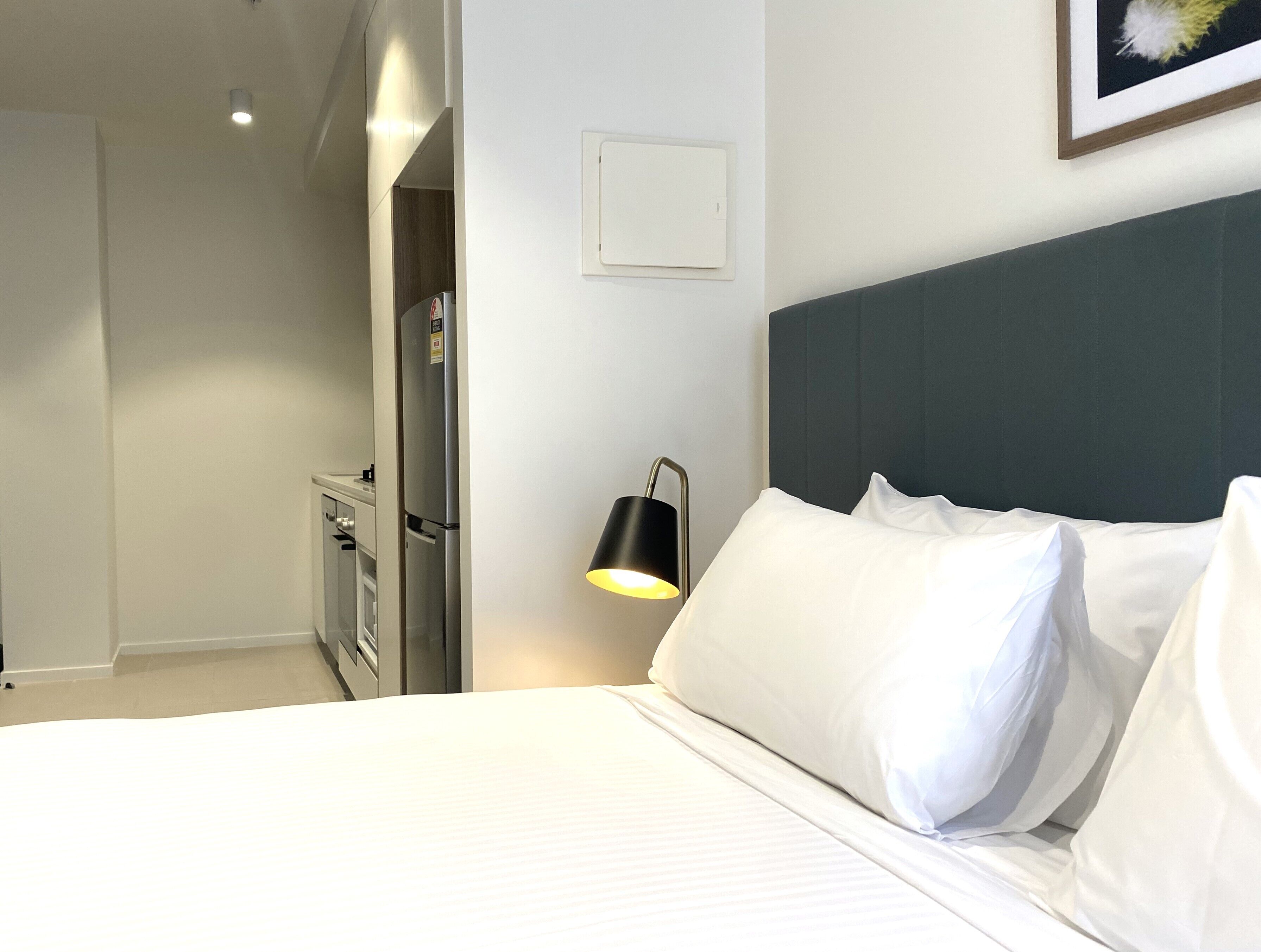 A hotel room with a bed, a nightstand, a lamp, a wall-mounted light switch, and a door leading to a bathroom.