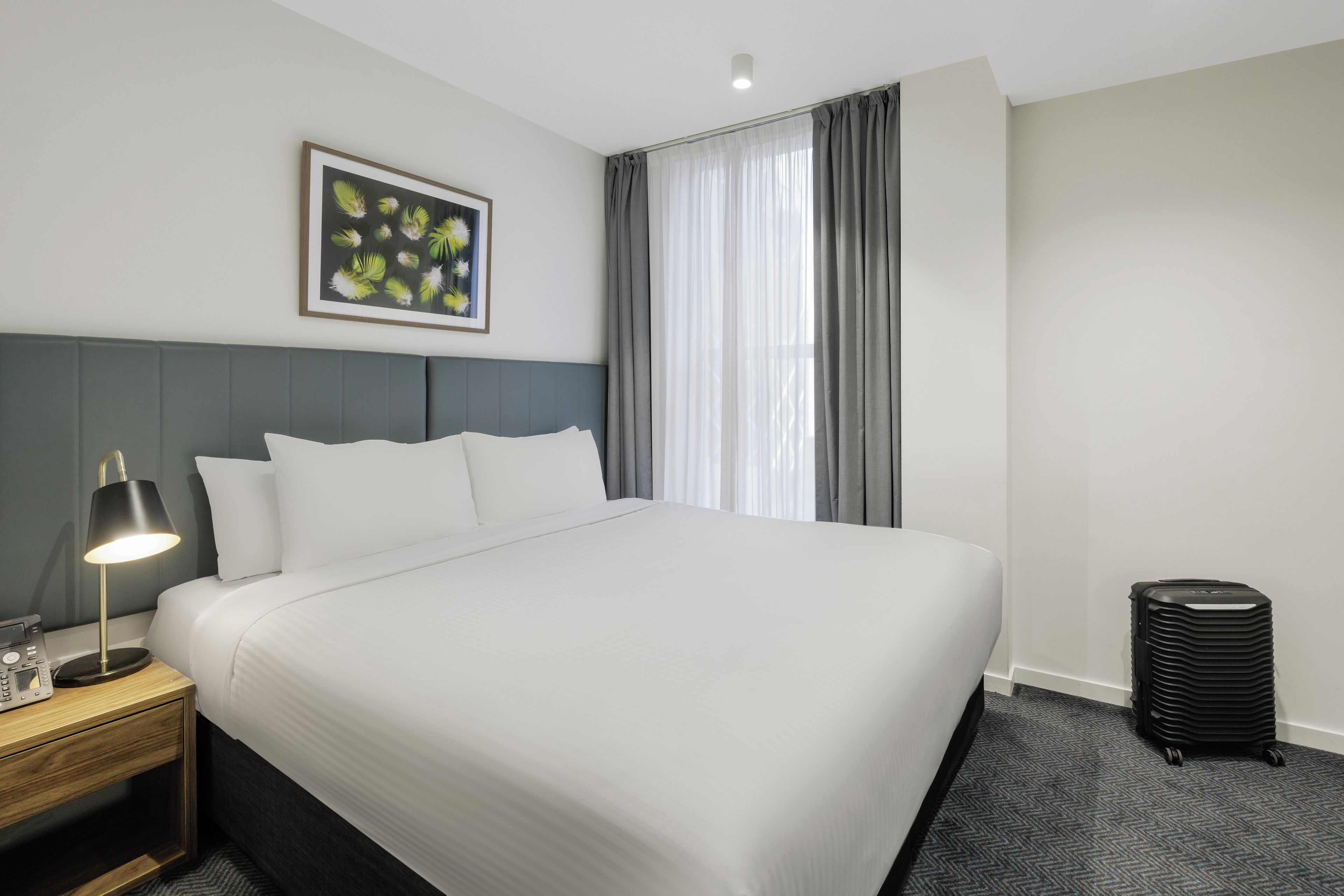 A hotel room with a large bed, a bedside table with a lamp, a framed picture on the wall, and a window with curtains.