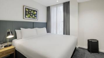 A hotel room with a large bed, a bedside table with a lamp, a framed picture on the wall, and a window with curtains.