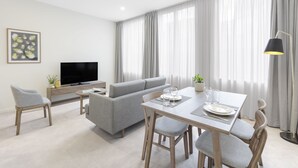 A modern living room with a grey sofa, a dining table with chairs, a flat-screen TV, and a large window with curtains. - Brady Apartment Hotel Hardware Lane (Melbourne)