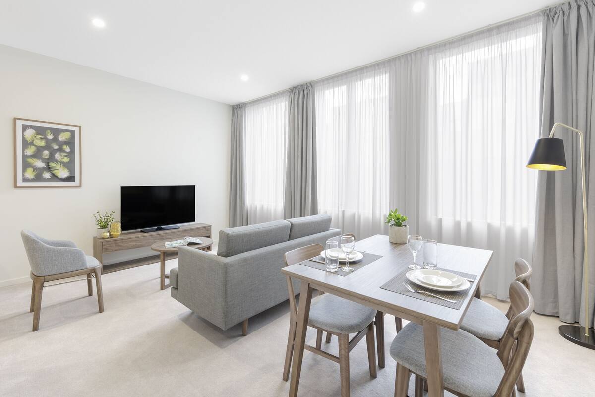 Una sala moderna con un sofá gris, una mesa de comedor con sillas, un televisor de pantalla plana y un ventanal con cortinas.