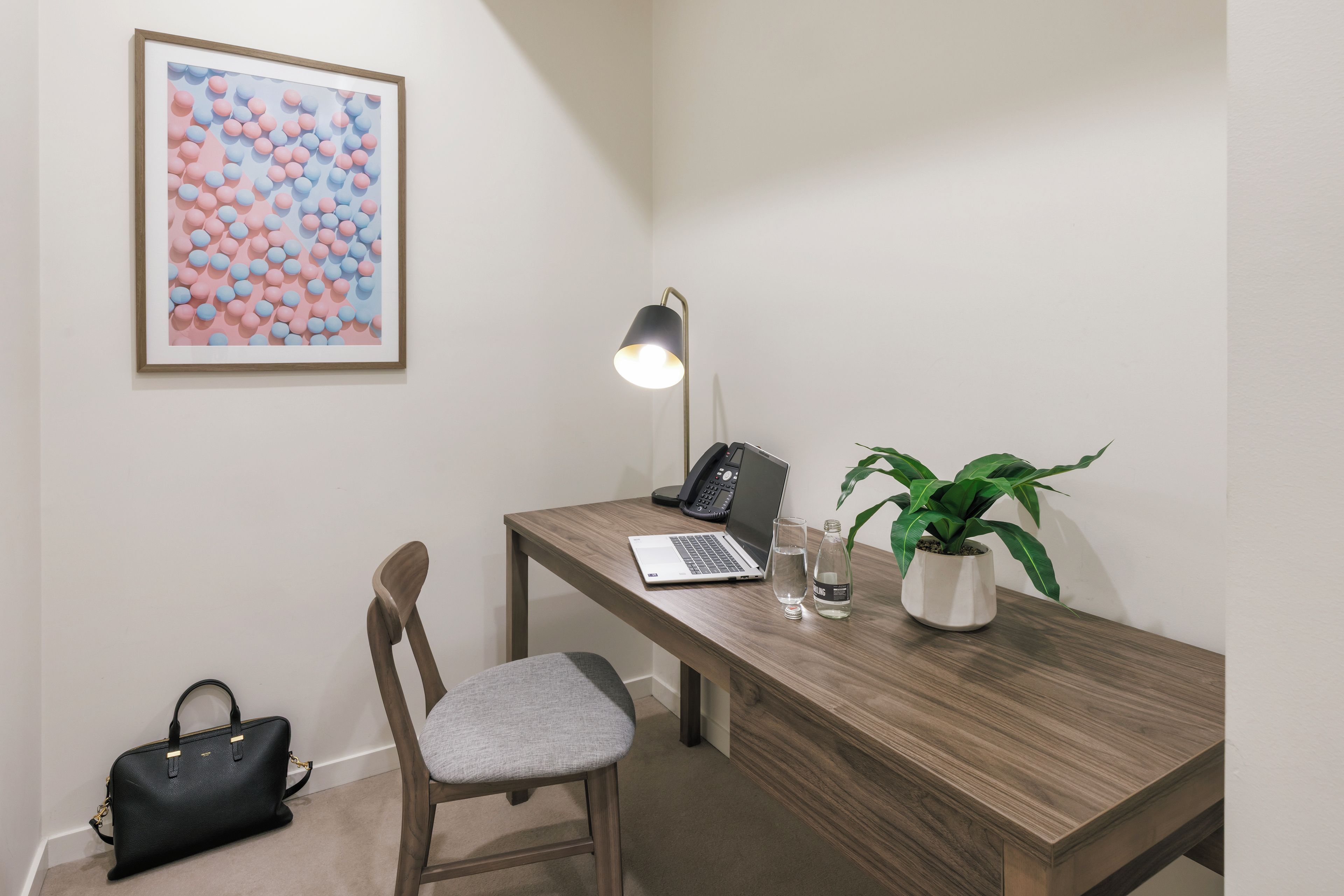 A workspace with a desk, chair, laptop, lamp, glass, bottle, and handbag.