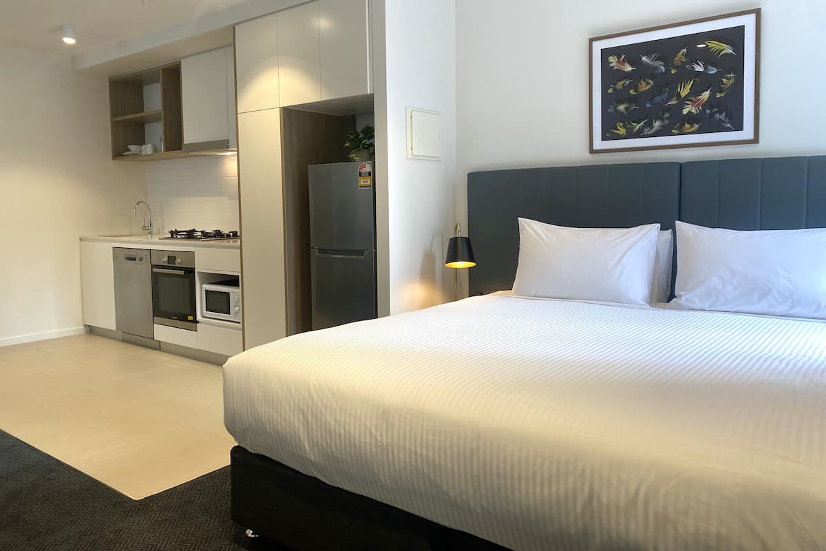 Una habitación de hotel moderna con cama, mesita de noche, un cuadro en la pared y una kitchenette con refrigerador y horno.