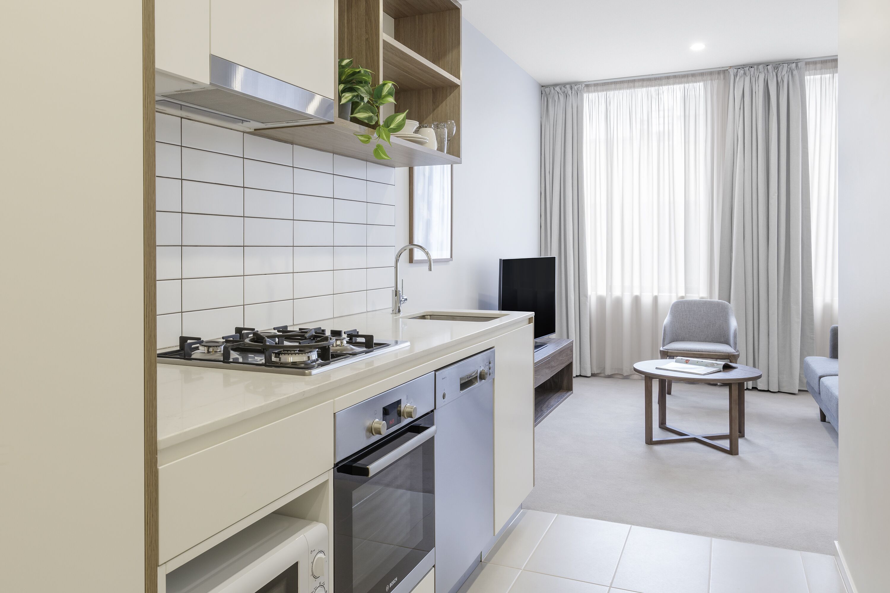 A modern kitchen with a gas stove, oven, and dishwasher, a dining area with a table and chair, and a large window with curtains.