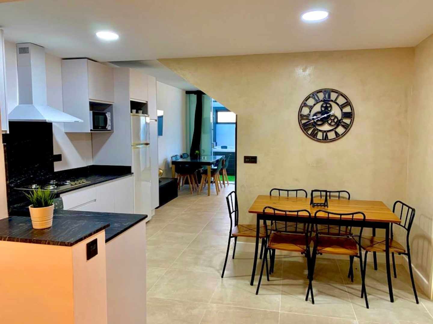 A modern kitchen with white cabinets, a black countertop, and a stove. There is a dining area with a wooden table and chairs, and a large wall clock.