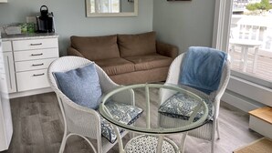 Una sala de estar acogedora con un sofá marrón, dos sillas de mimbre, una mesa de centro de vidrio y un aparador blanco con cajones.