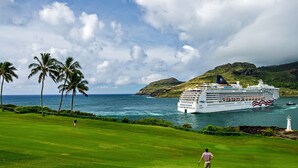 A cruise ship with multiple decks and a flag, sailing near a coastline with palm trees and a lighthouse. - Enjoy Perfect Vacation at Luxury Beach Front Resort. (Kapaʻa)