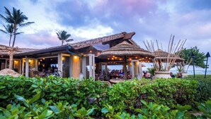 A beachfront restaurant with outdoor seating, palm trees, and a thatched-roof structure. - Enjoy Perfect Vacation at Luxury Beach Front Resort. (Kapaʻa)
