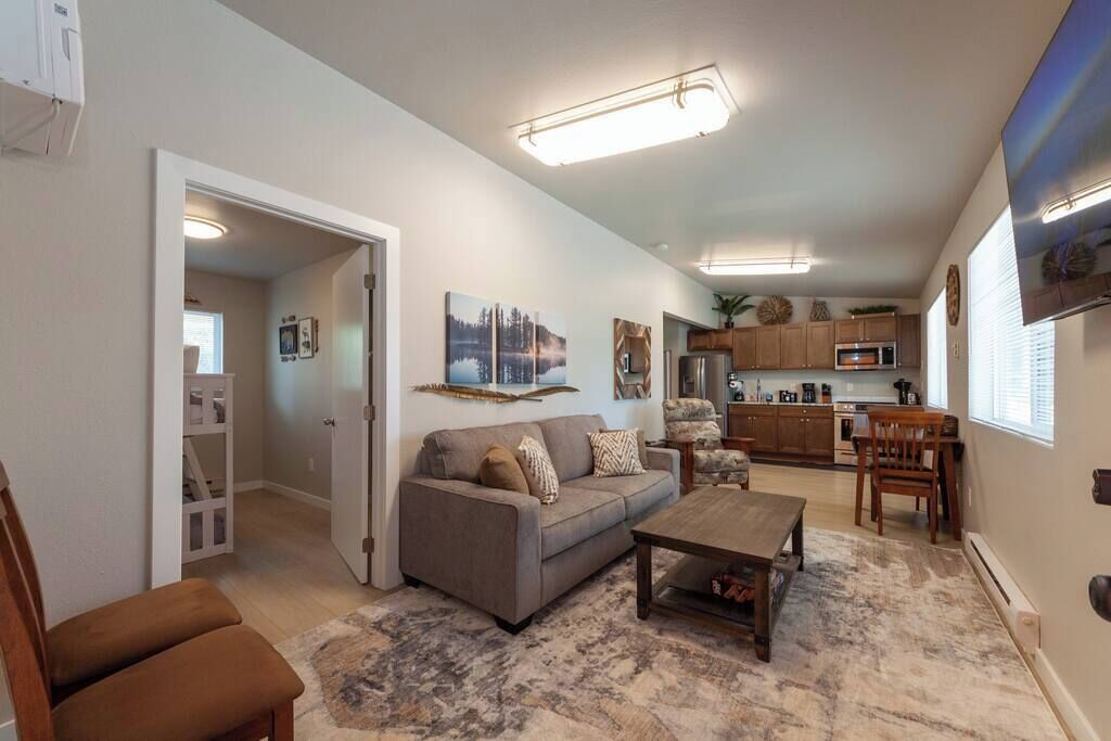 A living room with a sofa, coffee table, and a dining area with chairs. There is a kitchen in the background with cabinets and appliances.