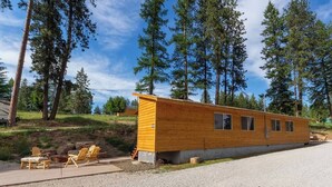 Una cabaña de madera con un pequeño porche y zona de estar al aire libre, rodeada de altos pinos.