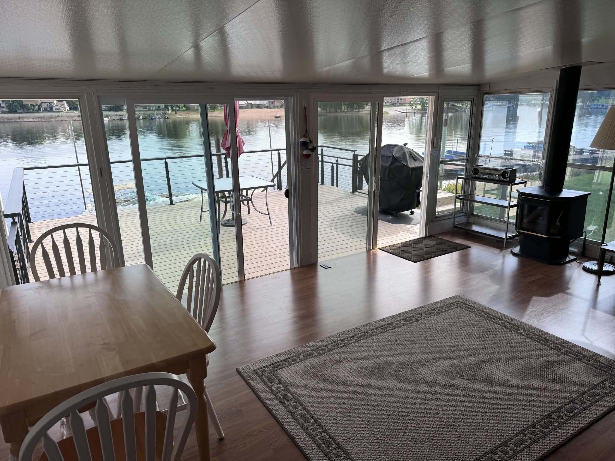 A room with a wooden table, chairs, a rug, a grill, and a view of a water body through large glass doors.