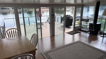 A room with a wooden table, chairs, a rug, a grill, and a view of a water body through large glass doors.