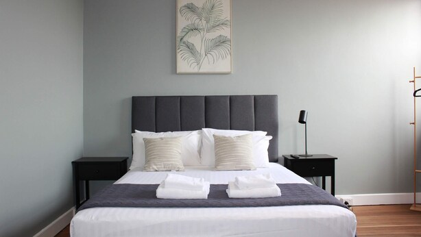A neatly made bed with white linens and two pillows, a bedside table with a lamp, and a framed botanical print on the wall. - Colliery Inn, Wallsend (Wallsend)