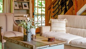 A cozy room with a wooden staircase, a sofa with a cushion, a wooden table with a vase and a candle, and a window with a view of greenery. - Cabin By The Creek! Inground Pool! Spa! (Dayton)