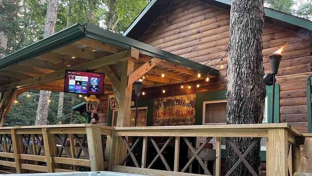 A wooden cabin with a hot tub, a TV screen, and a sign for plumbing and heating services. - Cabin By The Creek! Inground Pool! Spa! (Dayton)